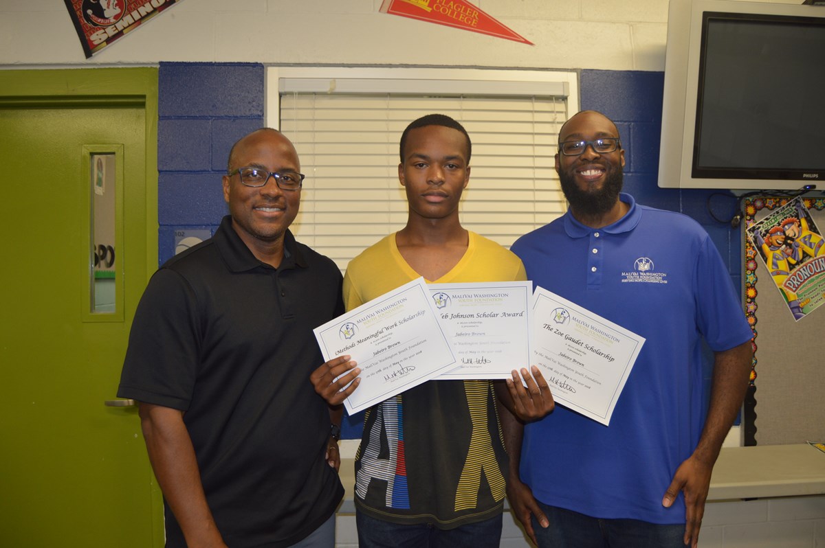 Jabeiro Brown holding his scholarship awards with two other men on either side of him in a classroom.